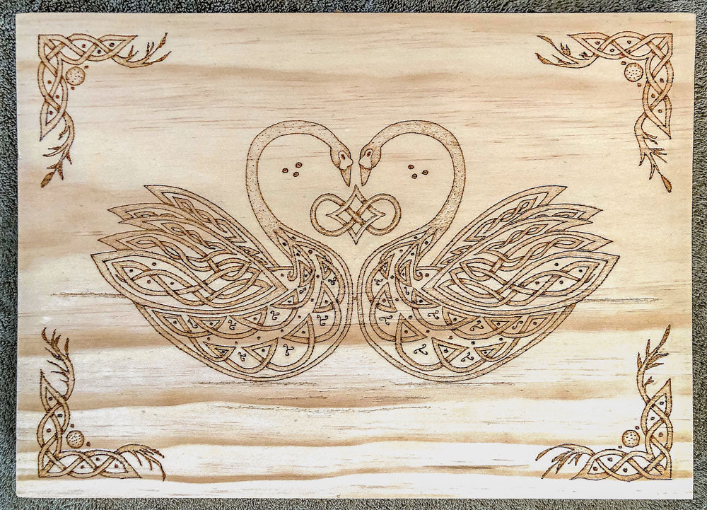 Wooden plaque with intricate Celtic knot design of two swans forming a heart shape.