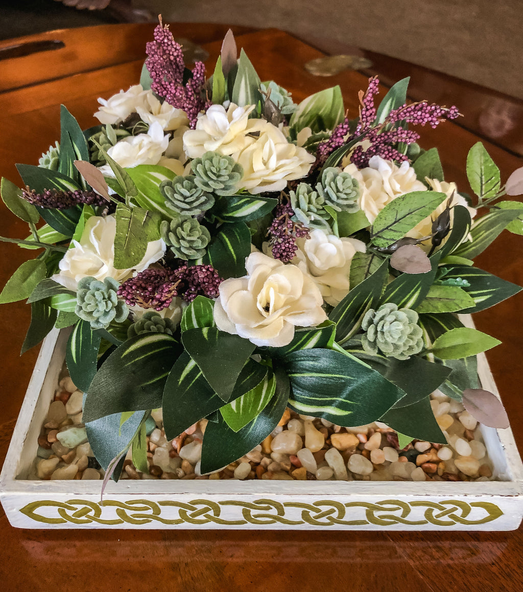 Faux floral arrangement with purple and white flowers and succulents in a gravel filled tray on a table