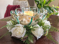 Candleholder with a striped glass bowl ringed with faux white roses and ferns on a table