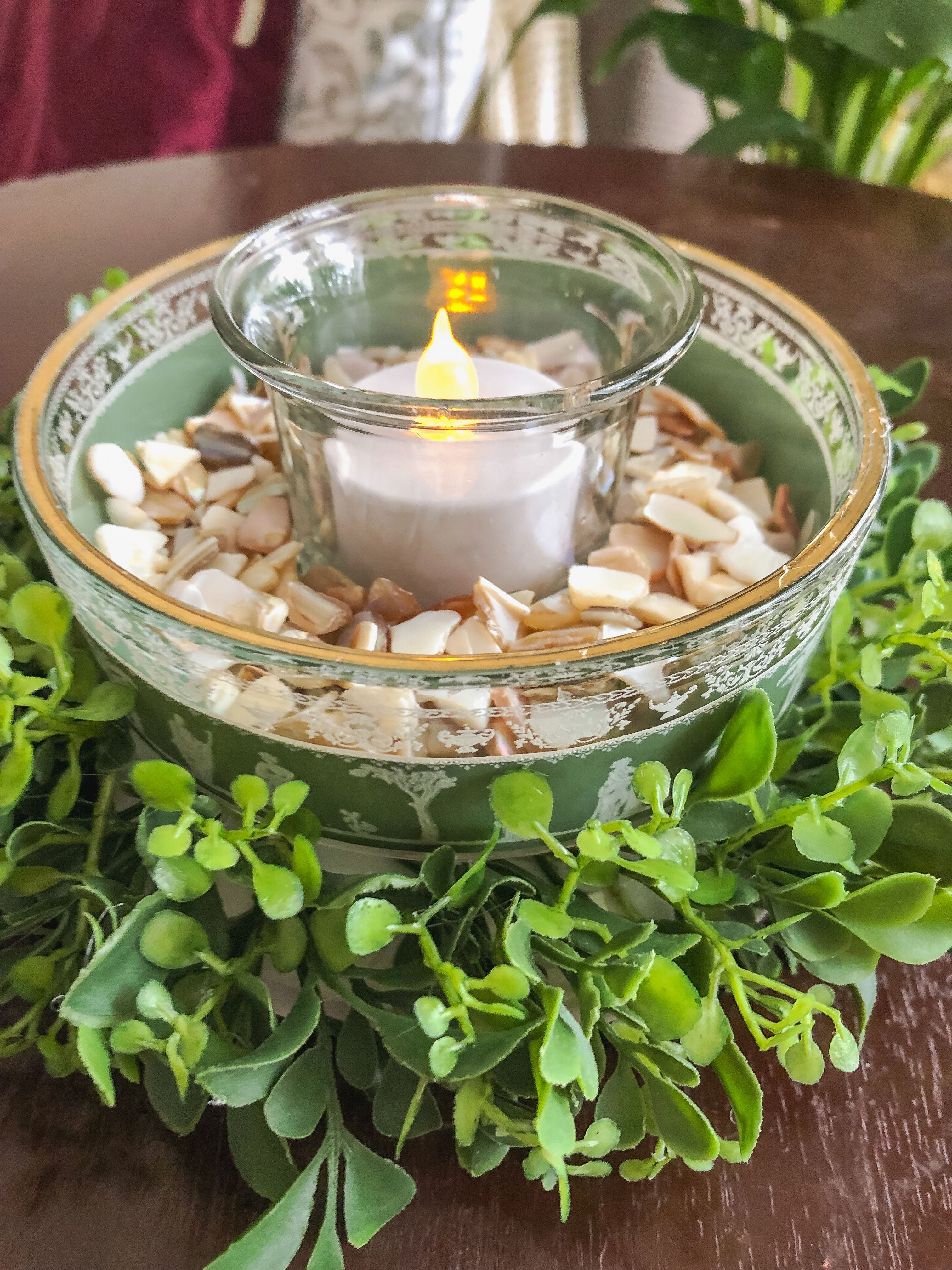 Candleholder with vintage glass filled with shell bits and ringed with faux boxwood on a table