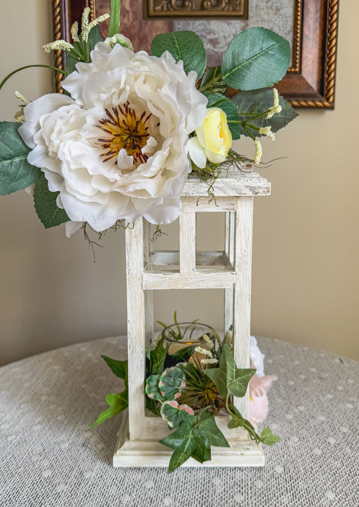 White lantern candleholder with faux summer florals on a lace table